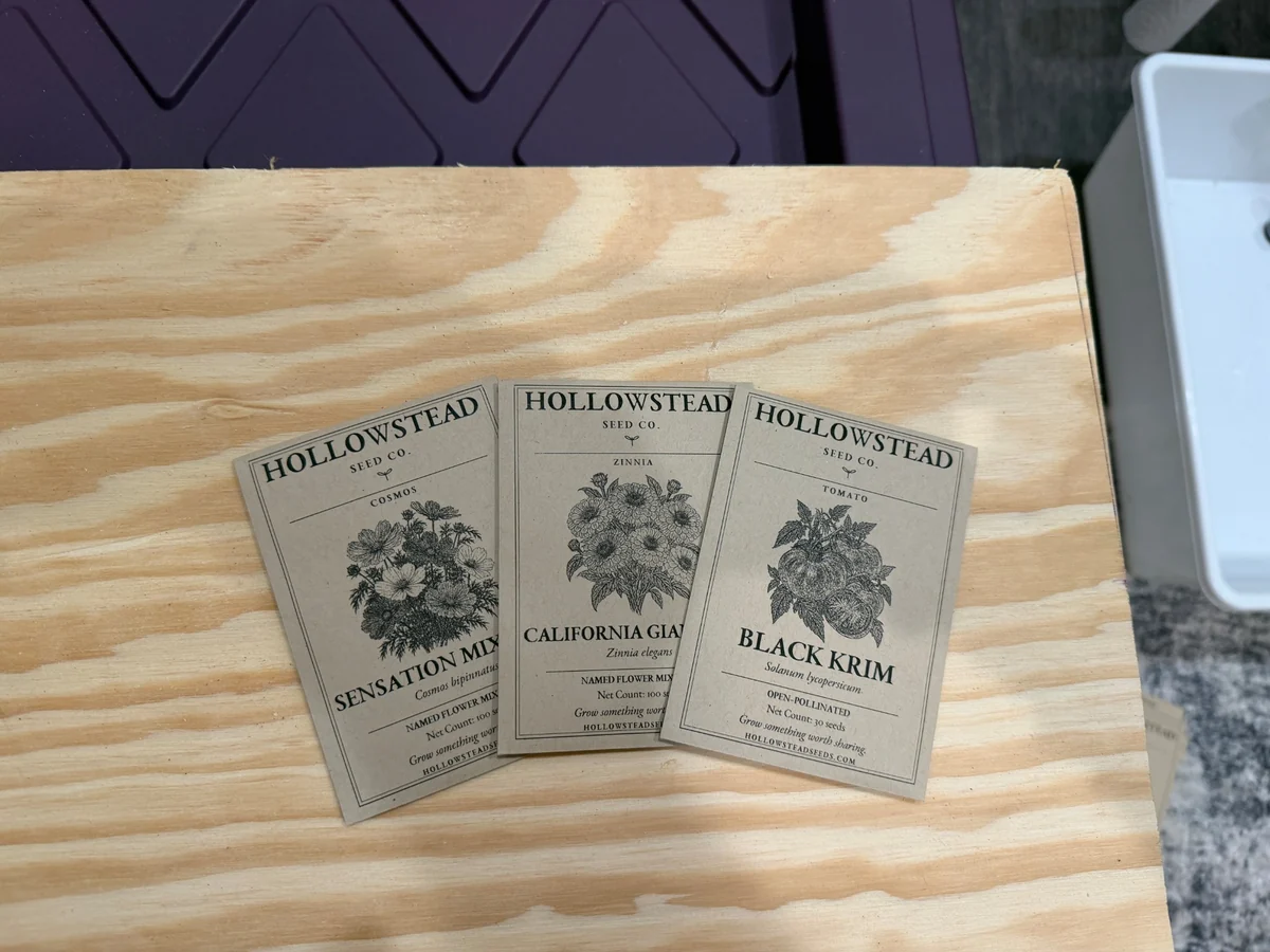 Hollowstead kraft seed packets for Sensation Mixture cosmos, California Giant zinnia, and Black Krim tomato on a wooden packing table.